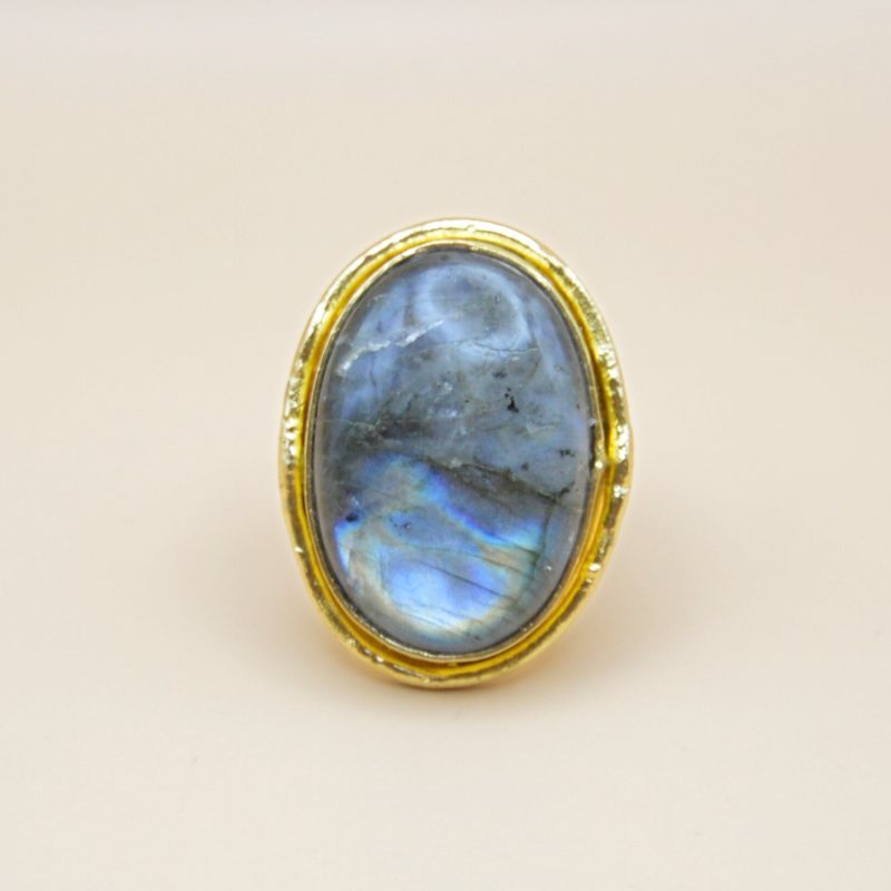 Confectionnée avec soin en laiton doré à l'or fin 18 carats, la bague Mystique est réglable et capture l'essence de l'inconnu et de la fascination. En son cœur, un imposant cabochon de labradorite scelle l'alliance entre l'éclat doré et la profondeur changeante de la pierre. Bijou seul, Crédit GEMITY