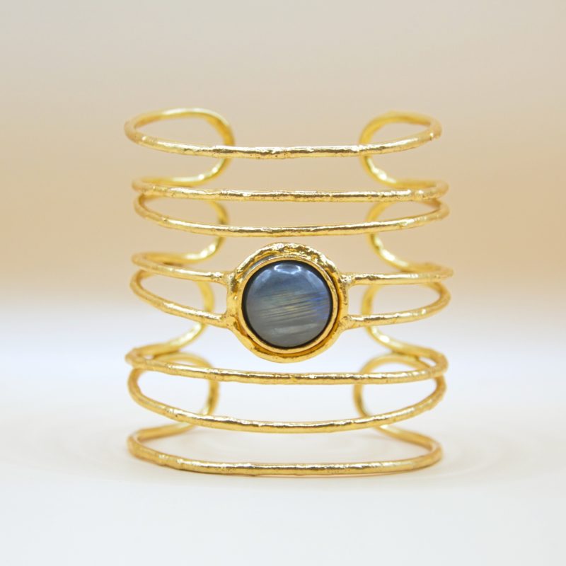 la manchette Kiera en laiton doré à l'or fin incarne l’élégance moderne avec une pierre de labradorite ronde, resplendissante au centre. Sa silhouette large et ajourée offre un mélange audacieux de sophistication et d’originalité. Bijou seul, Crédit GEMITY