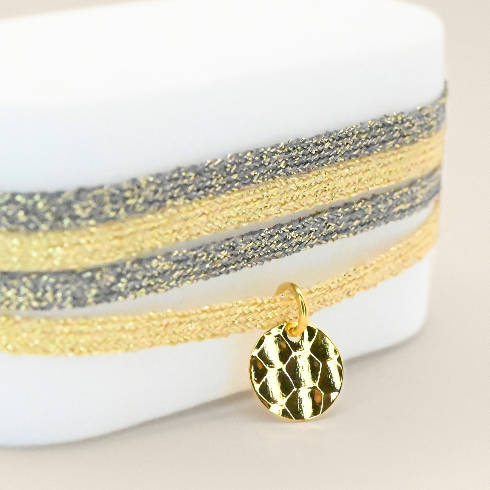 "Bracelet double tours en tissu de deux coloris différents avec une pastille martelée dorée à l’or fin. Fermoir en plaqué or réglable . Bijou seul, Crédit GEMITY"