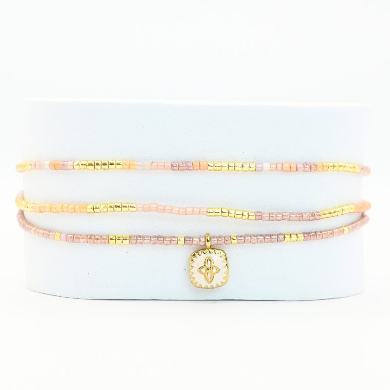 ce bracelet fin à 3 rangs combine des perles dorées et nude pour un effet élégant et doux. Il est orné d'un charm fleur doré, posé sur un fond en nacre blanc en forme de carré, apportant une touche de sophistication et de brillance. . Bijou seul, Crédit GEMITY