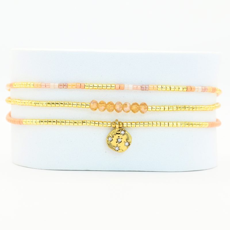 ce bracelet fin à 3 rangs combine des perles dorées, nude et roses, créant une harmonie de couleurs douce et élégante. Le charm rond à strass, placé au centre, ajoute une touche scintillante et raffinée. . Bijou seul, Crédit GEMITY