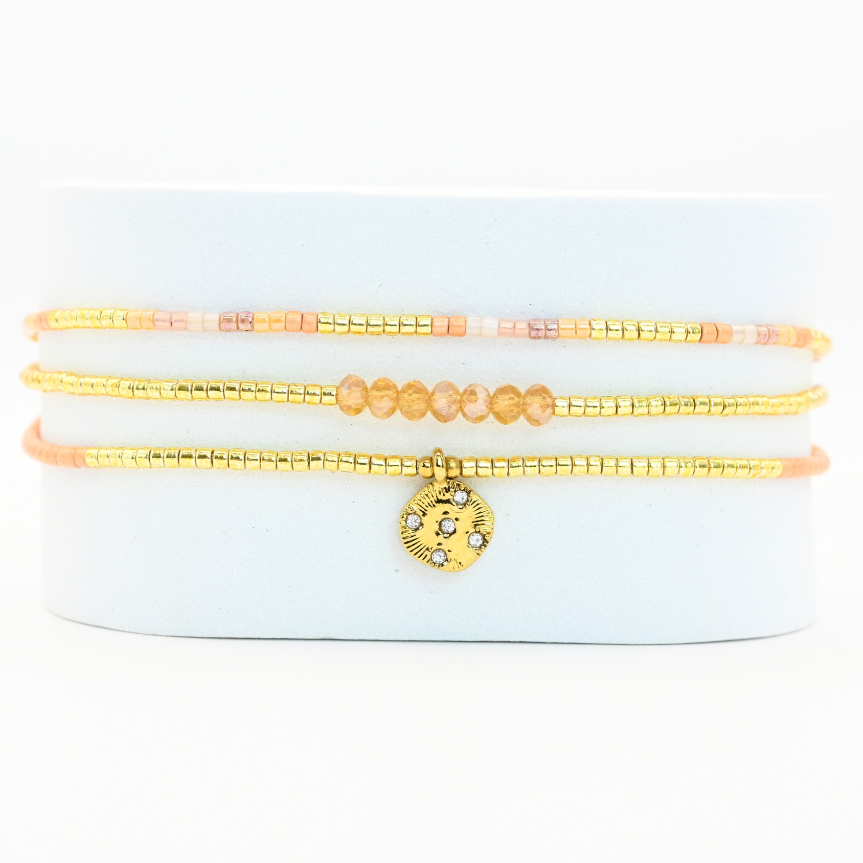ce bracelet fin à 3 rangs combine des perles dorées, nude et roses, créant une harmonie de couleurs douce et élégante. Le charm rond à strass, placé au centre, ajoute une touche scintillante et raffinée. . Bijou seul, Crédit GEMITY