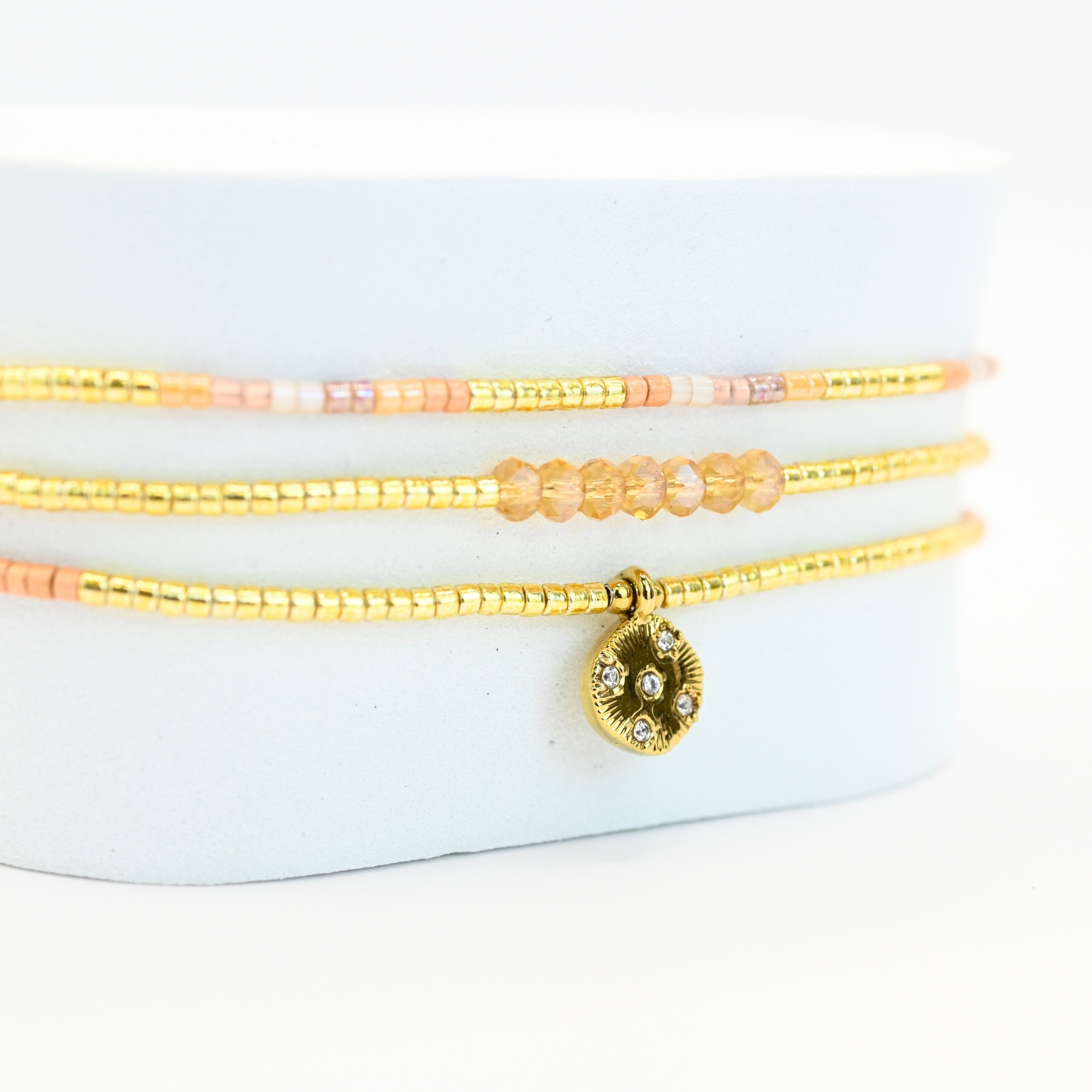 ce bracelet fin à 3 rangs combine des perles dorées, nude et roses, créant une harmonie de couleurs douce et élégante. Le charm rond à strass, placé au centre, ajoute une touche scintillante et raffinée. . Bijou seul, Crédit GEMITY