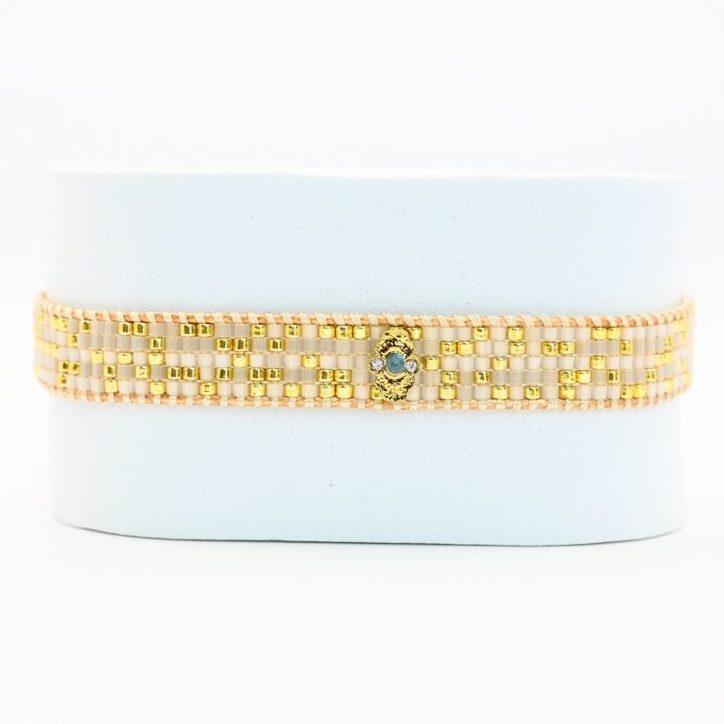 ajoutez une touche de sophistication à votre poignet avec cette petite manchette composée de 5 rangs de perles dorées et blanches. L'ornement doré en son centre apporte une touche de brillance et d'élégance supplémentaire à ce bijou délicat.. Bijou seul, Crédit GEMITY