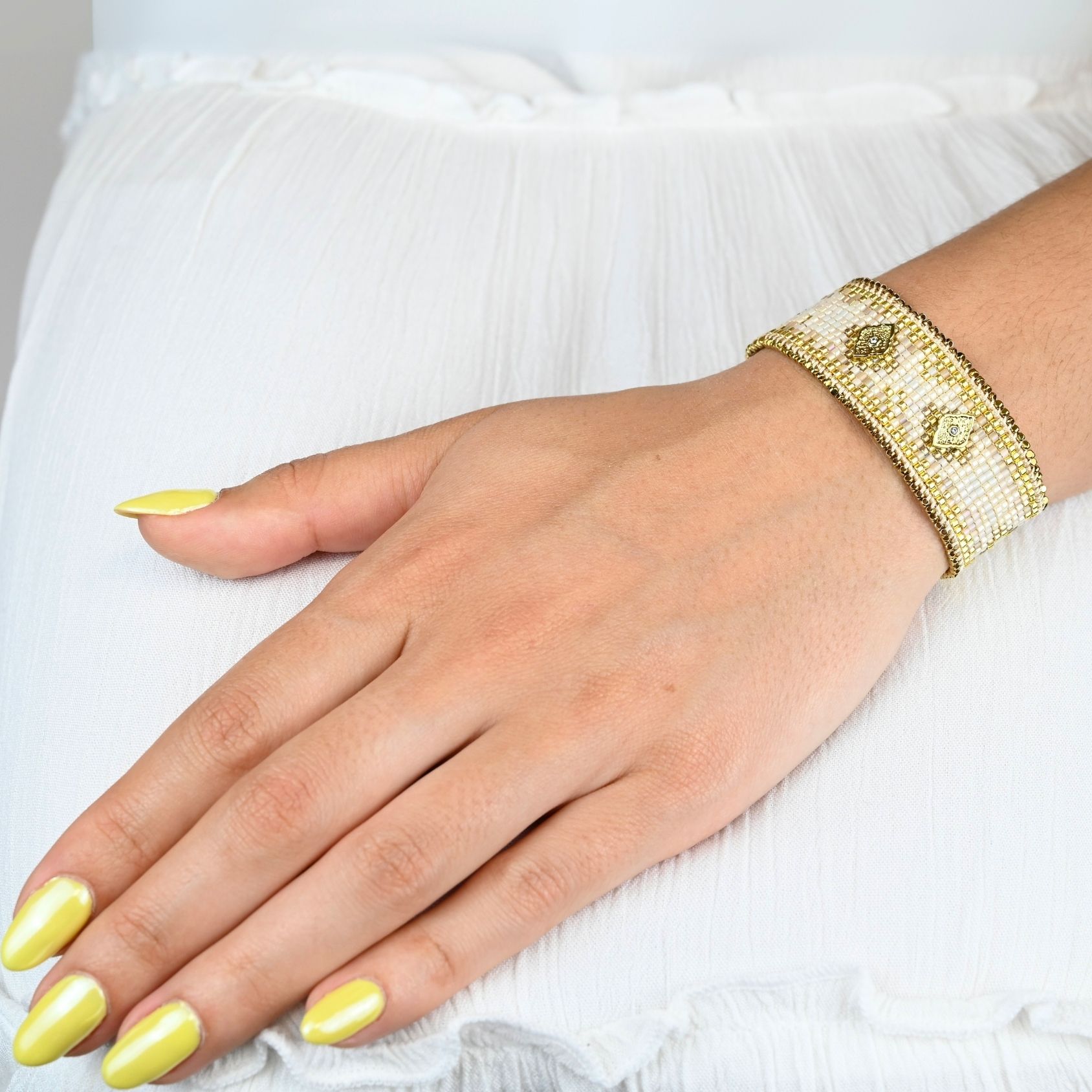 sublimez votre poignet avec cette grande manchette composée de 12 rangs de perles dorées et nude, créant un contraste élégant et lumineux. Deux ornements losange, placés au centre et à chaque extrémité, ajoutent une touche géométrique moderne et raffinée. Bijou porté, Crédit GEMITY.